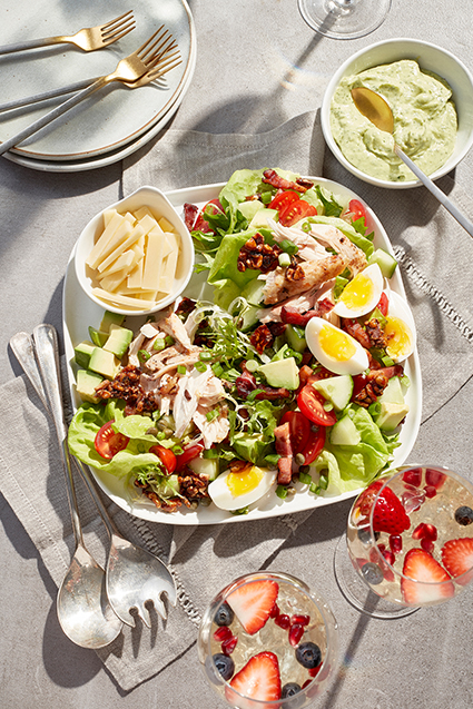 A colorful brunch salad made of mixed greens, sliced eggs, avocado cubes, tomatoes with a small bowl of sliced cheese.