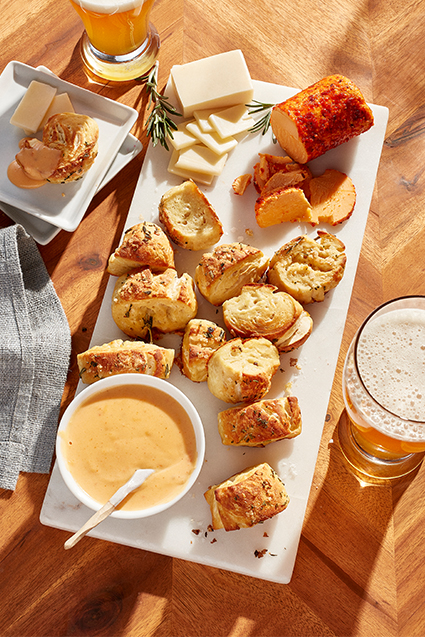 A board filled with cheesy herb pretzel bites, cheese logs and dips.