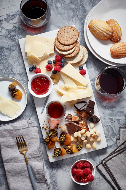 A charcuterie dessert board filled with slices of cheddar cheese, dark chocolate, nuts, jam, blueberries, raspberries and crackers.