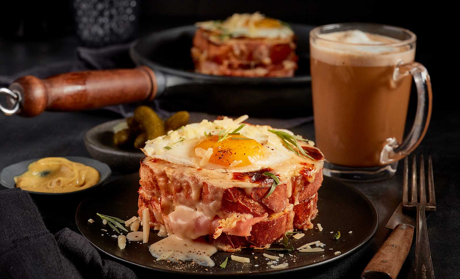 An up-close shot of a croque madam with a cappuccino and pickles in the background.