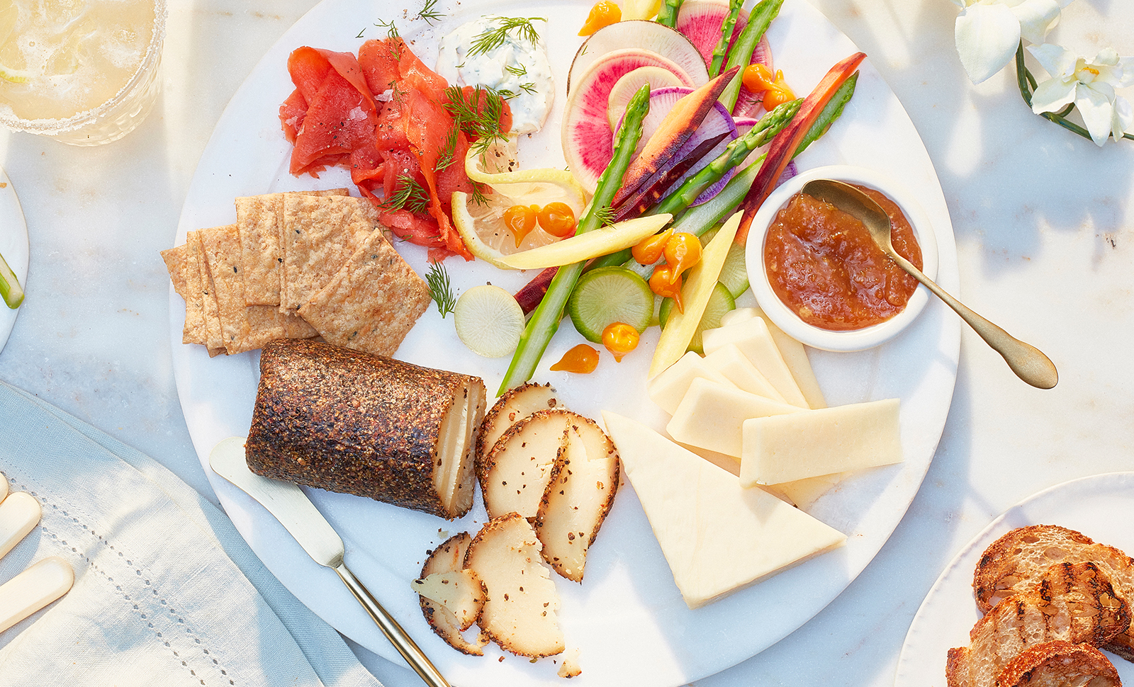 A spring inspired charcuterie board with a cheese log, cheddar cheese slices, crackers, tomatoes, cucumbers and asparagus.