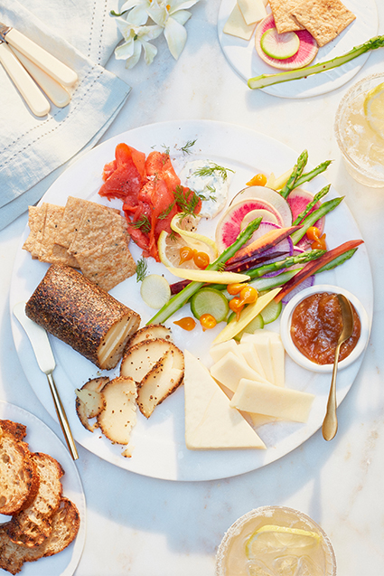 A spring inspired charcuterie board with a cheese log, cheddar cheese slices, crackers, tomatoes, cucumbers and asparagus.
