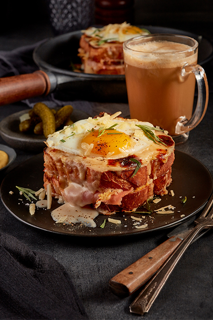 An up-close shot of a croque madam with a cappuccino and pickles in the background.