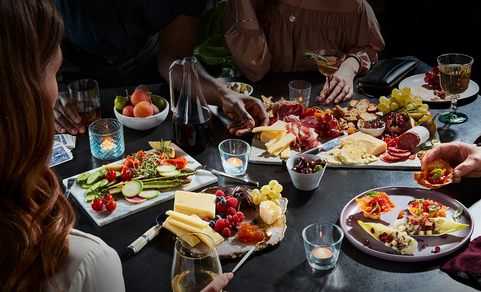 Four people around a table dressed in several platters of assorted cheeses, meats, fruits, vegetables, crackers, nuts, and cocktails.