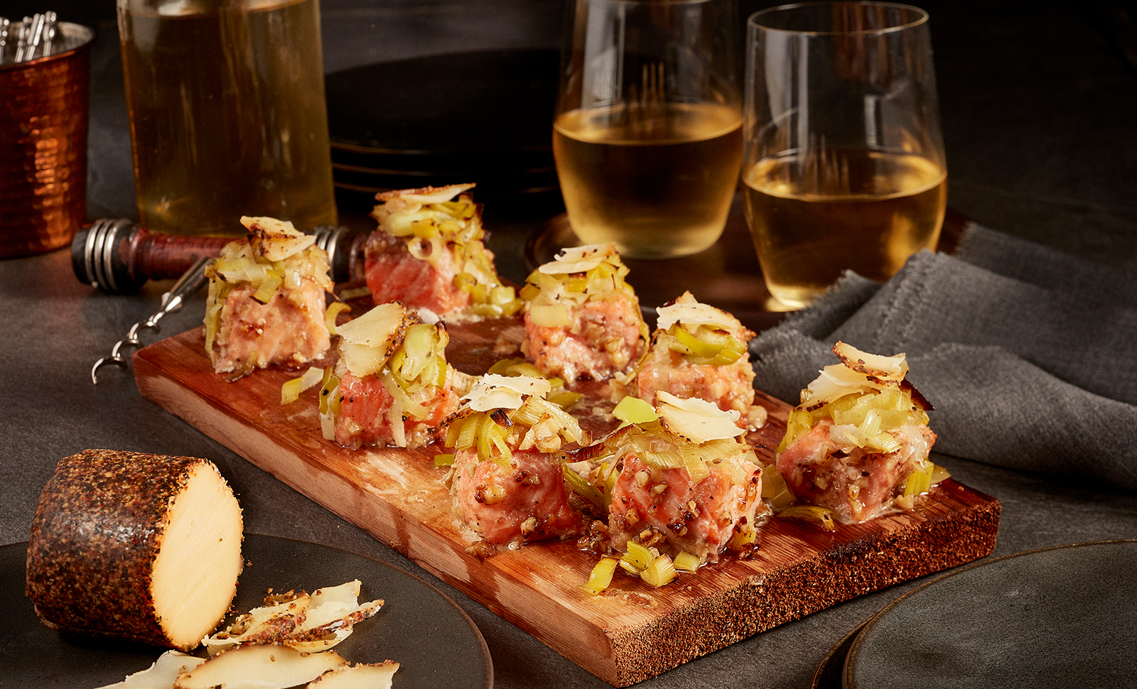 Maple walnut salmon bites served on a cedar plank with two glasses of white wine and Maple Cheddar with cracked black pepper cheese log.