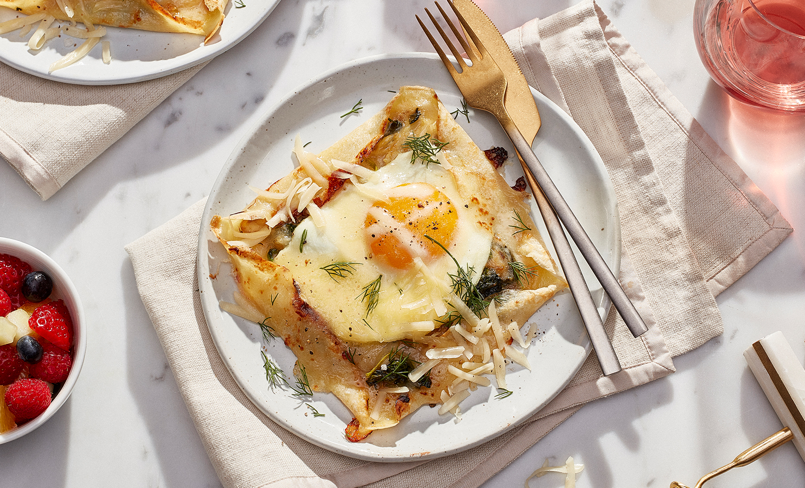 Open face savoury cheddar herbs crepe on a white plate with a fork and knife next to a small bowl of mixed fruits.