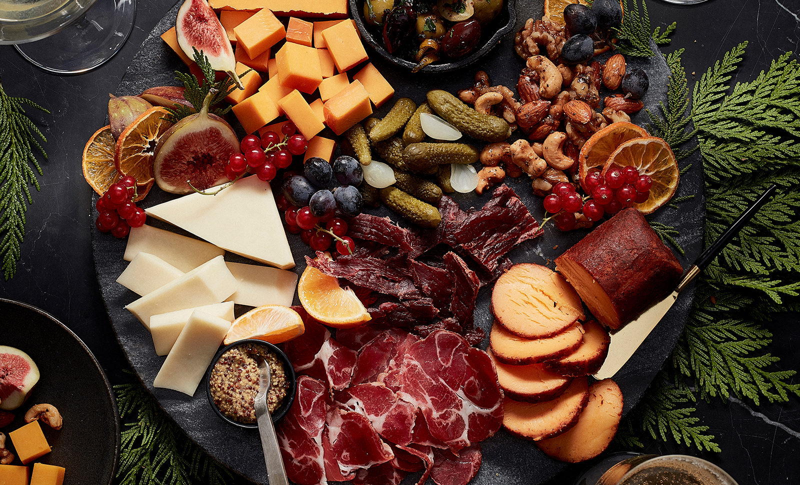 A winter inspired charcuterie board with a chipotle seasoned cheese log, cheddar cheese cubes, sweet pickles, mixed raw nuts, orange slices, grapes, blueberries and sliced meat.