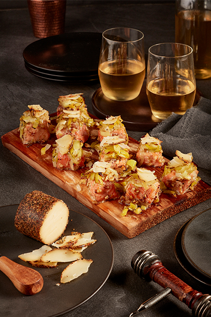 Maple walnut salmon bites served on a cedar plank with two glasses of white wine and Maple Cheddar with cracked black pepper cheese log.